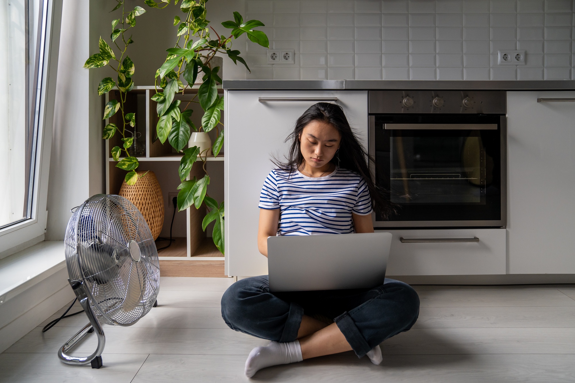 Handsome asian girl student with laptop is languishing heat sitting on cool floor next fan.